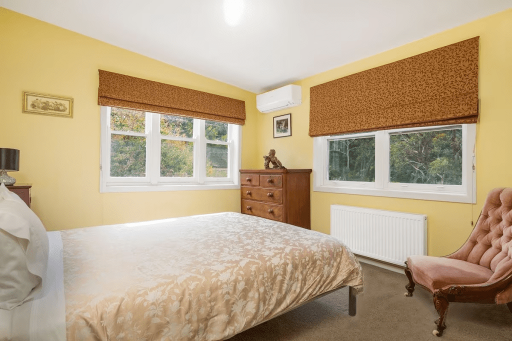 Bedroom with yellow walls, queen bed, gold bedspread, two windows looking out to gardens, heater and air-con.